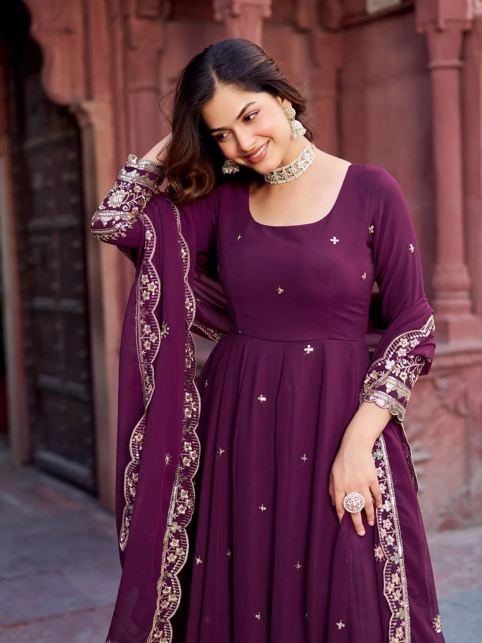 A woman stands gracefully in a deep plum-colored embroidered ethnic gown with a matching dupatta.